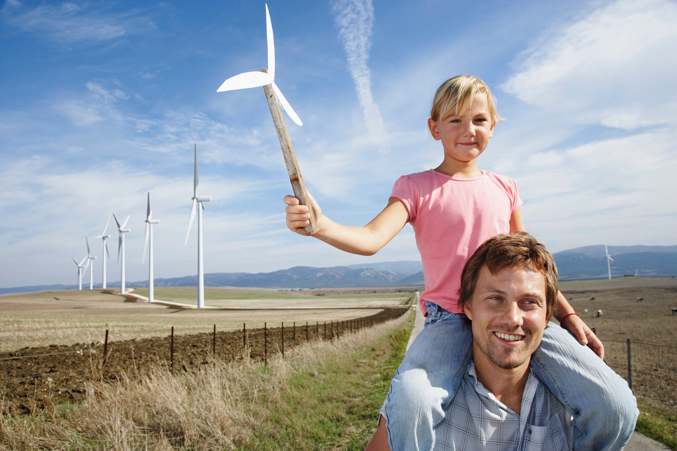 Daughter on father's shoulder holding a pinwheel standing in front of wind farm 
