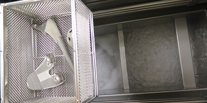 Close-up of orthopedic implants and parts in a vapor degreaser basket above bubbling Novec 73DE fluid.
